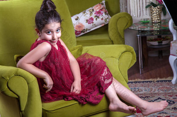 可爱的中东女孩，穿着<strong>深红</strong>色的连衣裙，收集头发，摆姿势，躺在家里的绿色沙发上。