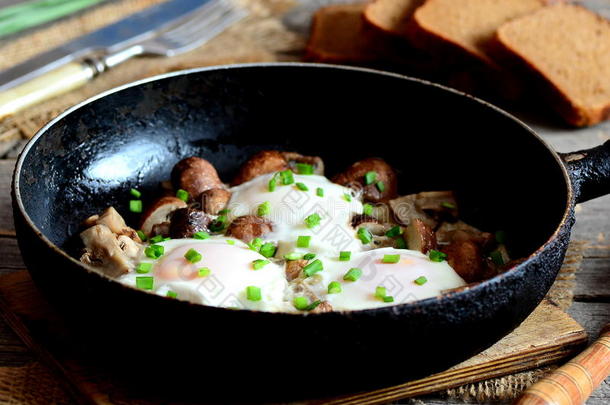 煎鸡蛋和蘑菇在煎锅，棕色面包片，叉子和刀在木制桌子上。 美味而简单的鸡蛋早餐