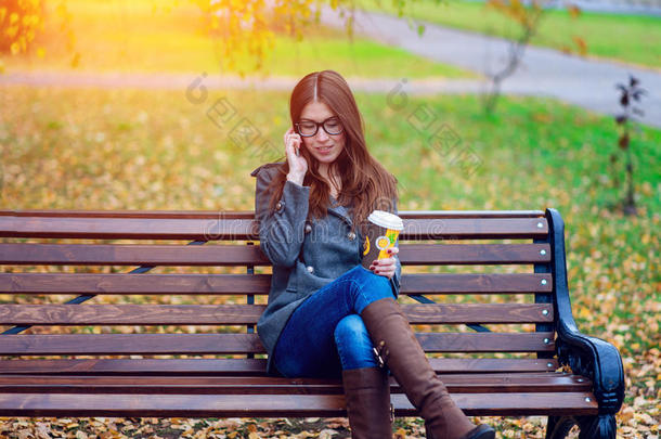 穿着夹克的女孩打电话，坐在长凳上，拿着咖啡或茶，年轻的户外，春天的秋天，生活