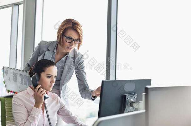 年轻的女商人用固定电话，同事指着办公室的<strong>电脑显示</strong>器