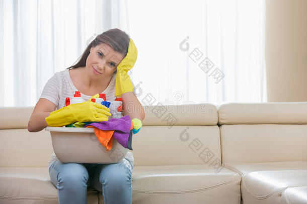 一位疲惫的妇女拿着一篮子清洁用品坐在家里的沙发上