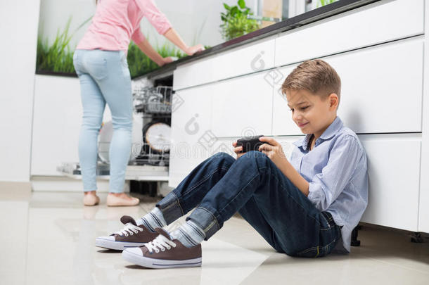 男孩在厨房里用手持式电子游戏机，背景是母亲