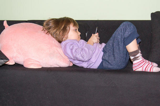 可爱的小女孩躺在床上，抱着她的玩具猪和手机