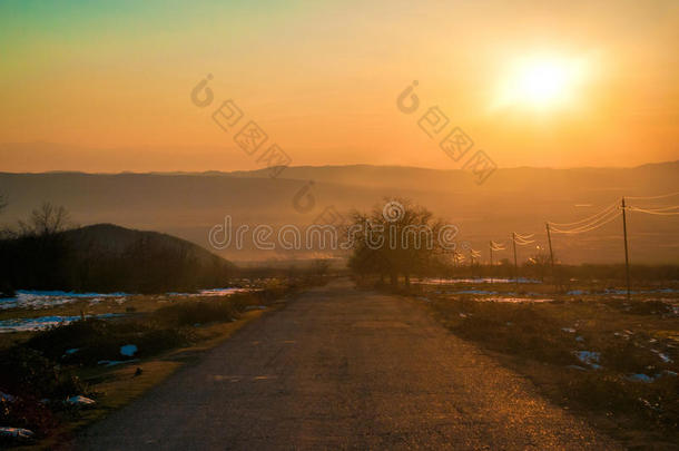 秋天或冬天的景观与道路和树木。 金色的光束日落。 在群山和天空的背景上有云。