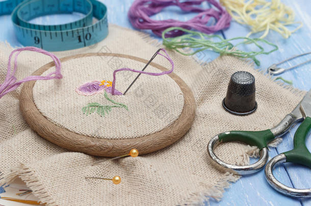 在木制刺绣框架中的特写线和亚麻织物，用于刺绣、剪刀、顶针和蓝色木制缝纫表