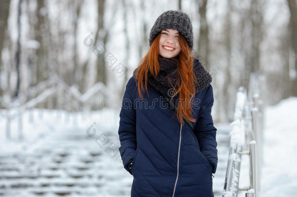 美丽的冬天肖像，年轻<strong>可爱</strong>的红头发女人戴着<strong>可爱</strong>的针织帽子，冬天在雪地公园的楼梯上玩得很开<strong>心</strong>