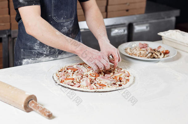 做披萨。 在面团<strong>预制</strong>件上安排肉类成分。 厨师面包师的特写手穿着制服的蓝色围裙