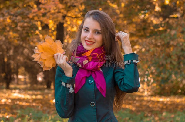 穿着粉红色围巾的漂亮微笑的女孩站在公园里，保留着树叶