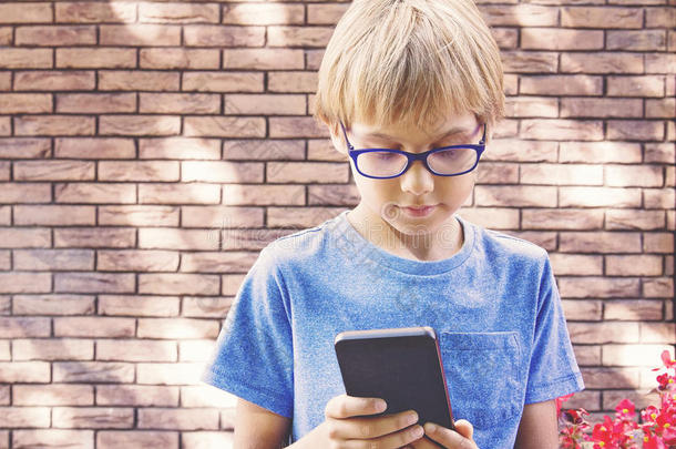 孩子在户外拿着手机。戴眼镜的男孩看着智能手机屏幕，使用应用程序，玩。砖墙
