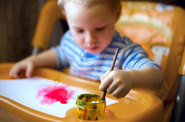 一个小男孩坐在孩子们的桌子旁，拿着画笔，油漆，油漆