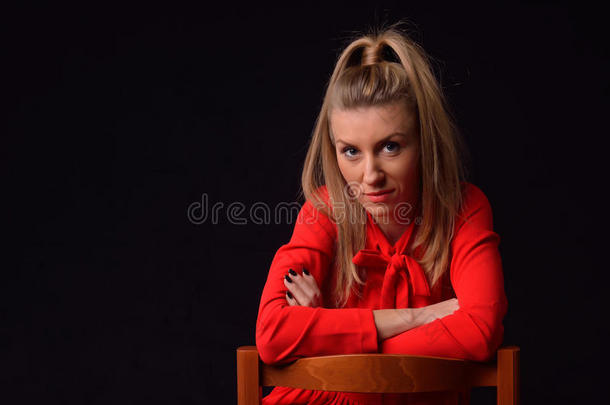 穿着红色连衣裙的漂亮金发年轻女人坐在<strong>一把椅子</strong>上，在黑色背景的工作室里摆姿势