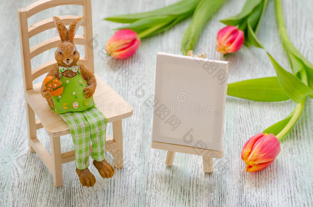 复活节兔子坐在凳子上，拿着一个鸡蛋，在木制背景上<strong>画画</strong>画架和郁金香