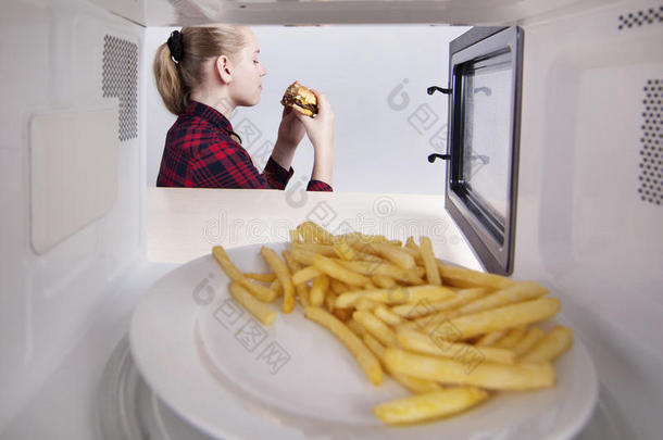 手里拿着汉堡包的女孩坐在桌子旁。 在微波炉里热一顿快餐。 通过烤箱查看