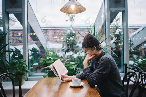 一个年轻的学生戴着帽子和衬衫，在读一本书，在一个美丽的咖啡馆和音乐学院喝咖啡