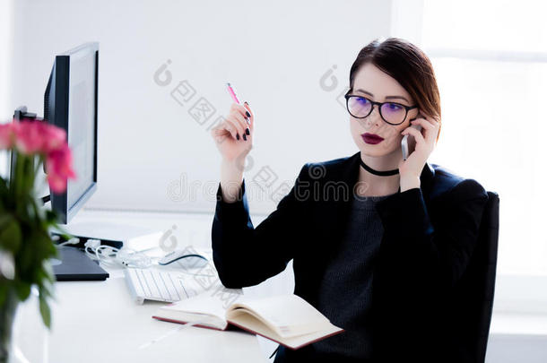 美丽的年轻女人坐在桌子旁，在凉爽明亮的办公室里打电话