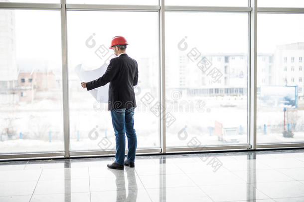 建筑工程师戴着安全头盔，看着全景窗户附近的蓝图纸施工