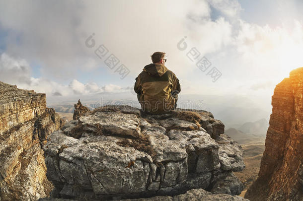 一个年轻人坐在一个由悬崖环绕的岩石上，位于<strong>云层</strong>上方的一个很大的位置，在<strong>云层</strong>的背景上