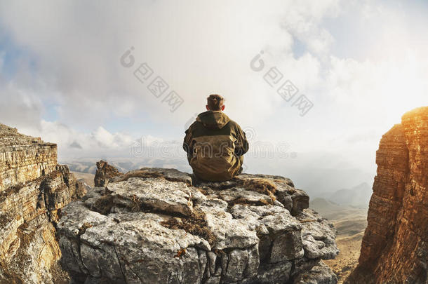 一个年轻人坐在一个由悬崖环绕的岩石上，位于<strong>云层</strong>上方的一个很大的位置，在<strong>云层</strong>的背景上