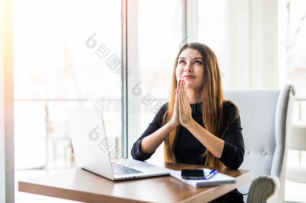 女商人祈祷。 穿着正式服装的体贴的女人，双手紧握，抬头看着笔记本电脑，一边坐着