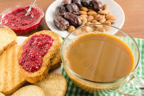 早餐时间，一杯加牛奶的咖啡，烤面包，覆盆子果酱，枣子，木桌上餐巾上的杏仁。