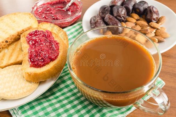 早餐时间，一杯加牛奶的咖啡，烤面包，覆盆子果酱，枣子，木桌上餐巾上的杏仁。