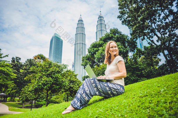 一位穿着休闲装的年轻女士在摩天大楼背景的热带公园里使用笔记本电脑。 移动办公概念