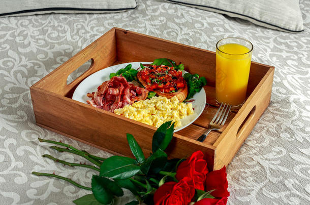 早餐在<strong>木托盘</strong>上放着一束红玫瑰。 碎鸡蛋，炸培根，面包和沙拉上的豆子，还有