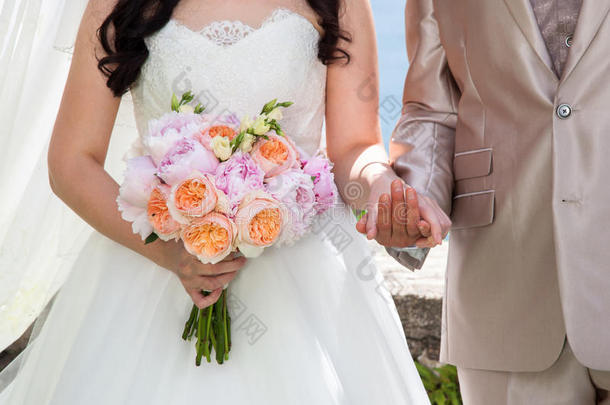 婚礼上的新娘和新郎用橙色玫瑰和粉红色牡丹的婚礼花束牵手。