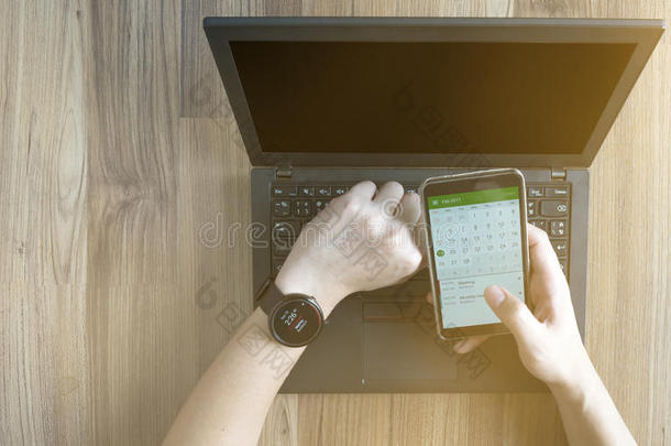 关闭男子检查时间表议程上的智能手表同步智能手机使用笔记本电脑在木桌上与阳光从窗口