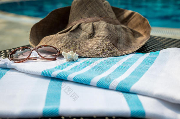 一条白色和蓝色的土耳其毛巾，太阳镜和草帽在藤椅上，背景是蓝色游泳池。