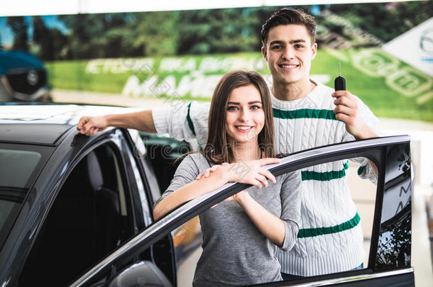 一对漂亮的年轻夫妇在车展上靠在他们的新车上，微笑着看着相机。 男人拿着车钥匙