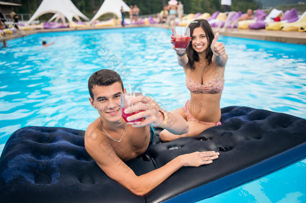 英俊的男人，有迷人的女人，在夏季度假胜地的游泳池里，在床垫上享用鸡尾酒