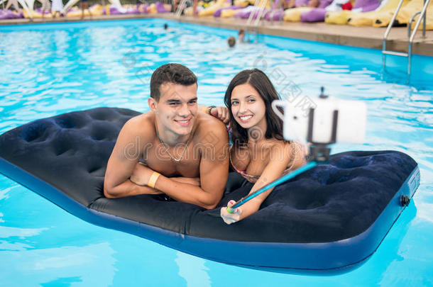 一对夫妇在手机上拍自拍照片，在游泳池和<strong>暑假的</strong>床垫上贴着自拍棒。