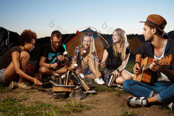 朋友们坐在篝火旁，微笑着，弹吉他。 野营烧烤棉花糖。