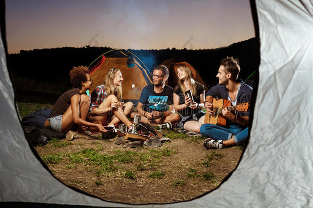 朋友们坐在篝火旁，微笑着，弹吉他。 野营烧烤棉花糖。