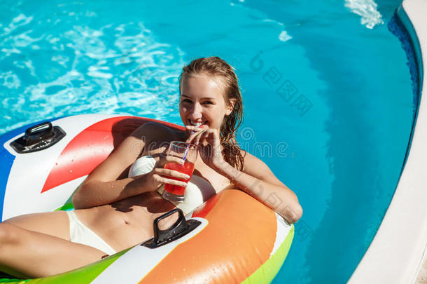美丽的金发女孩微笑着，喝着鸡尾酒，在游泳池里游泳。