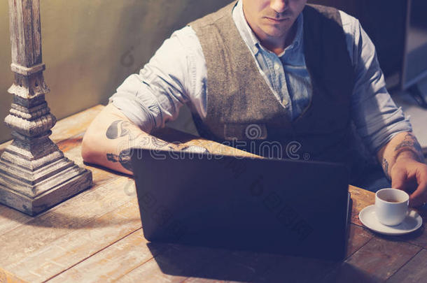 特写优雅的纹身男子在家工作，在笔记本电脑，而坐在木桌上。使用现代电脑