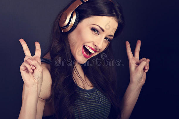 美丽快乐的年轻女子在无线耳机中听音乐，并在黑暗中用双手显示胜利的v-手势