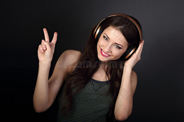 美丽快乐的年轻女子在金色无线耳机中听音乐，并在黑暗的GR上用手显示胜利的v-手势