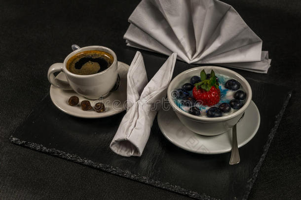 白色杯子里的黑色咖啡，甜点，大草莓和蓝莓的酸奶，在黑色的石板上，餐巾纸