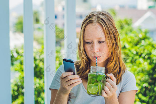 一个年轻的女人手里拿着一个梅森罐子，手里拿着一个莫吉托和一部智能<strong>手机</strong>。 <strong>春天</strong>的饮料概念