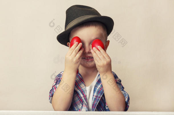 4岁的男孩戴着帽子和衬衫，双手捧着红色的复活节彩蛋，看着鸡蛋。