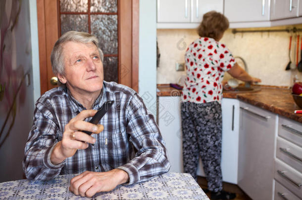 白种人的高级男人用电视遥控器切换频道，而坐在厨房的桌子旁，妻子洗餐具