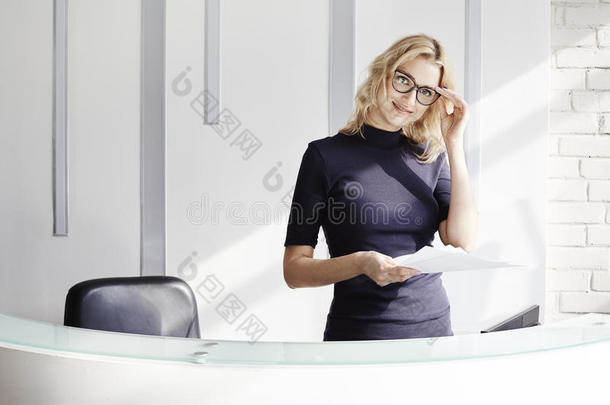 漂亮的金发友好的女人在<strong>接待处</strong>后面，管理员通过电话交谈。 现代办公室的阳光。