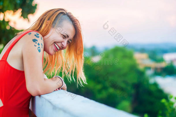 美丽的红发女孩穿着一件红色的衣服，在一家夏季餐厅的阳台上纹身笑着