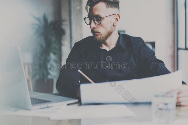 戴眼镜的大胡子年轻商人在现代办公室工作。男人使用当代笔记本电脑并做笔记