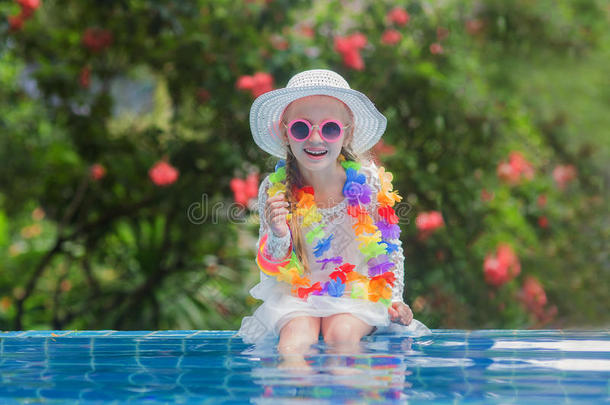 可爱的微笑女孩戴着太阳镜，花珠和一顶帽子，在游泳池里喝饮料