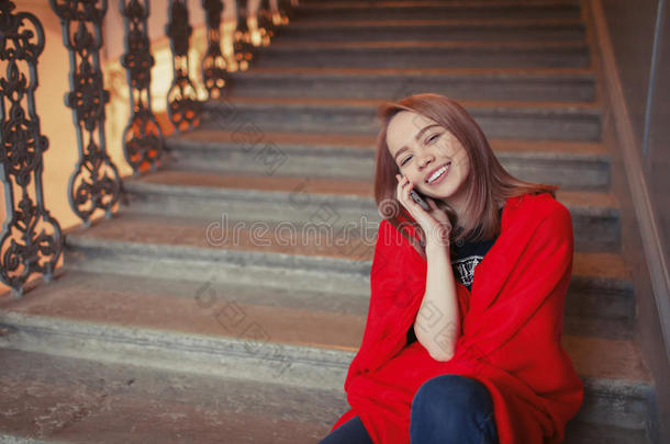 一个年轻的金发女孩坐在台阶上打电话，裹着一条红色的毯子。 她很快乐，微笑着