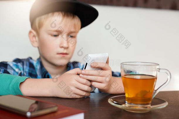 电影效果。 英俊的男孩穿着格子衬衫和帽子，用手机坐在咖啡馆里，喝了一杯茶，在智能p上输入了一条信息