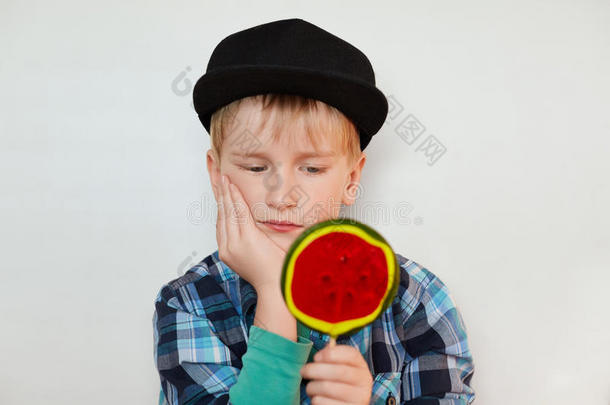 特写美丽的小男孩的肖像，戴着帽子和衬衫，一手拿着巨大的棒棒糖，严肃地看着它